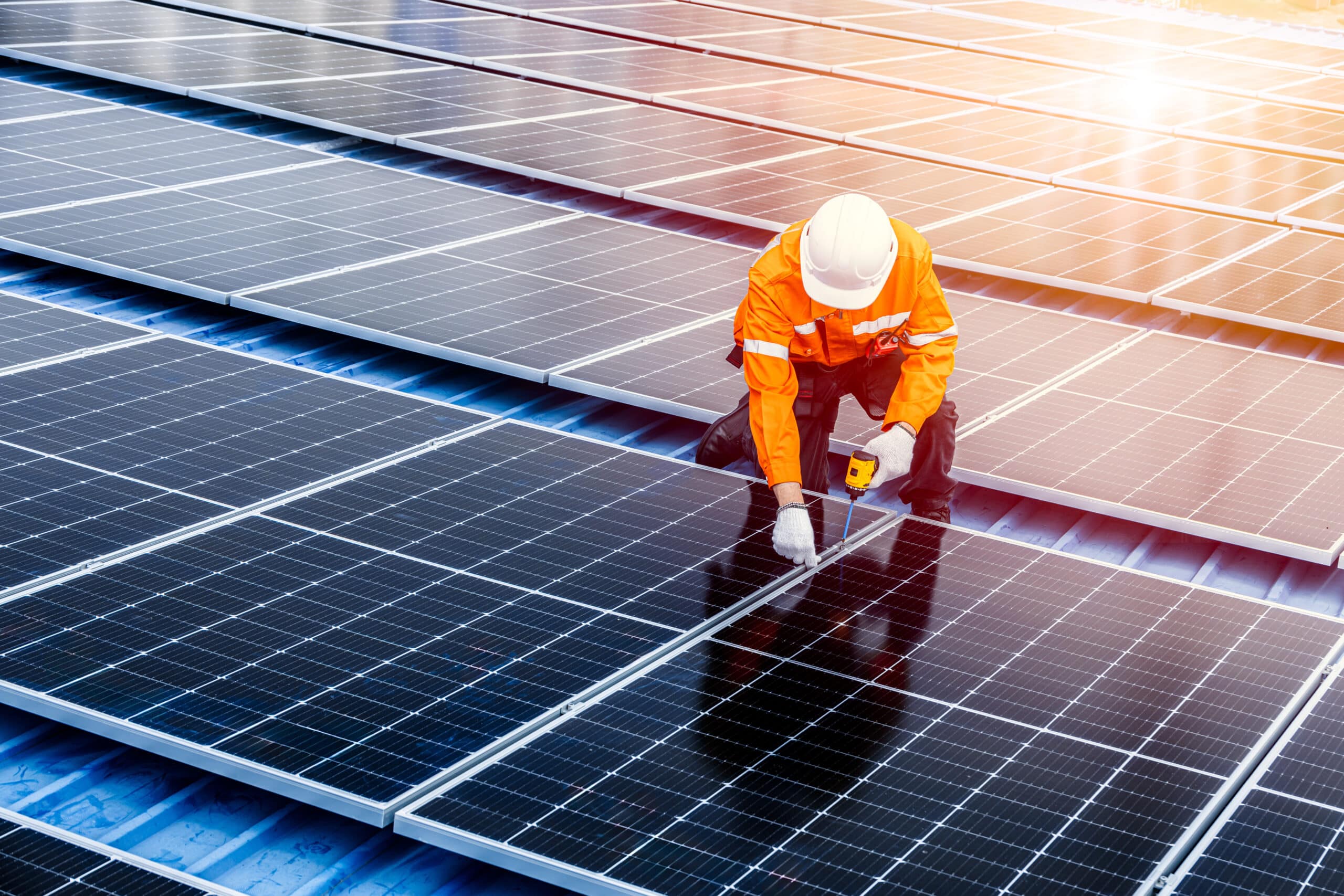 Technicien en combinaison orange installant des panneaux solaires sur un toit métallique bleu, sous un soleil éclatant.