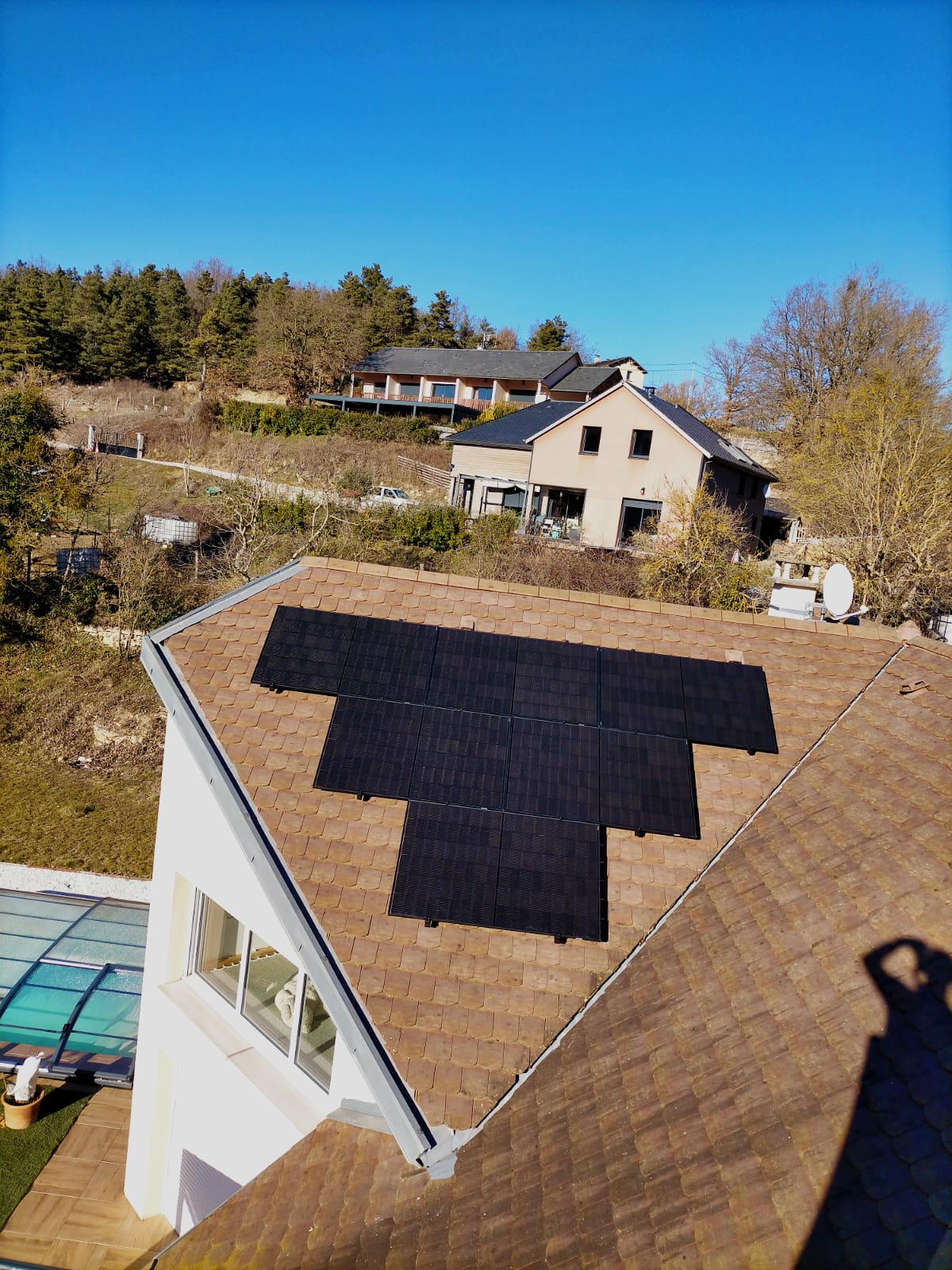 Installation de panneaux solaires noirs sur un toit de tuiles marron d'une maison sous un ciel bleu dégagé. Autres maisons et arbres en arrière-plan.