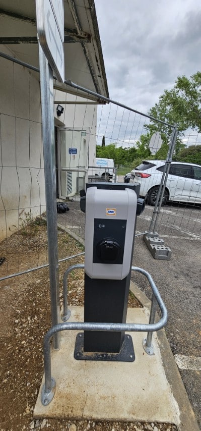 Borne de recharge VE DKV grise sur socle, avec barrières métalliques. Clôture de chantier, bâtiment, et parking avec SUV et van "OH GO" en arrière-plan.