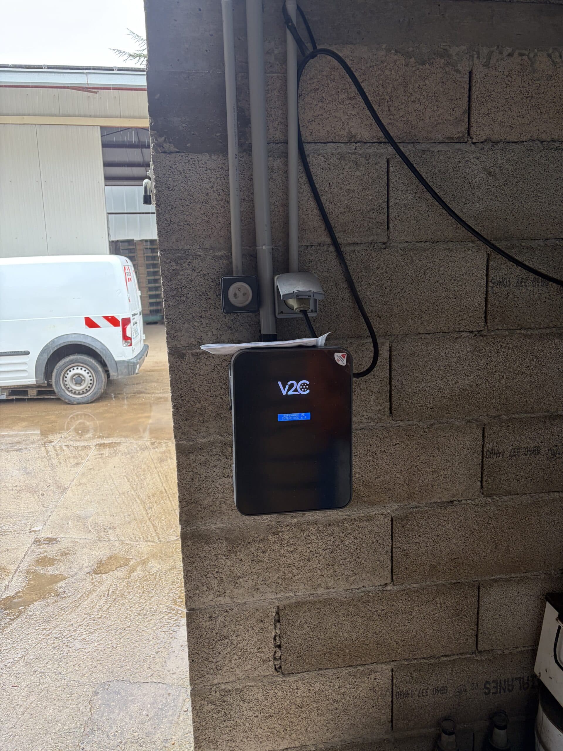 Borne de recharge électrique V2C sur mur en parpaings. Écran 'ESPERANDO VE'. Camionnette blanche à l'extérieur.