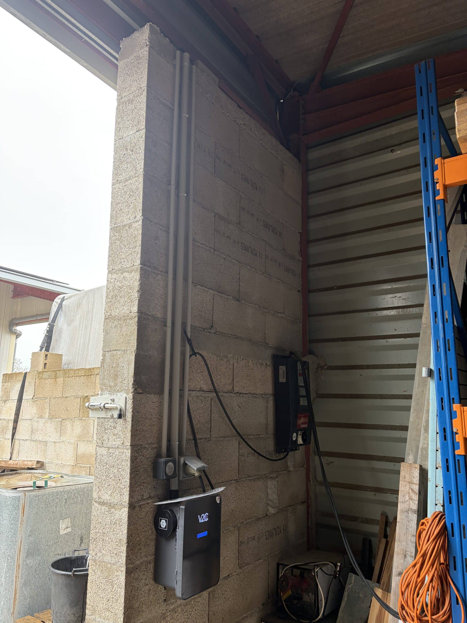 Borne de recharge V2C sur mur en parpaings dans un entrepôt avec conduits électriques et rack de stockage bleu.