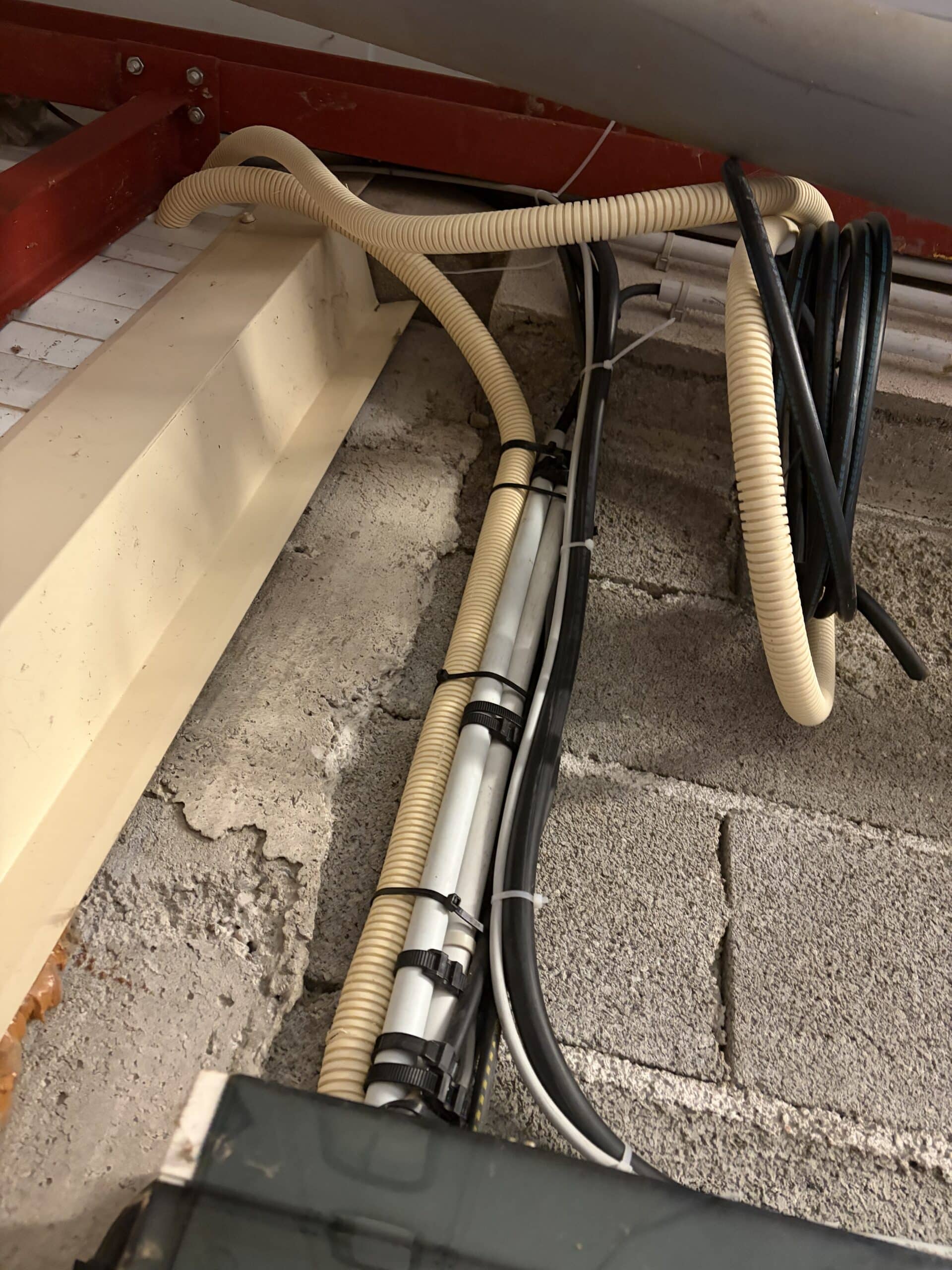 Tubes annelés, câbles électriques noirs et tuyaux PVC fixés par des serre-câbles sur un mur en parpaings.