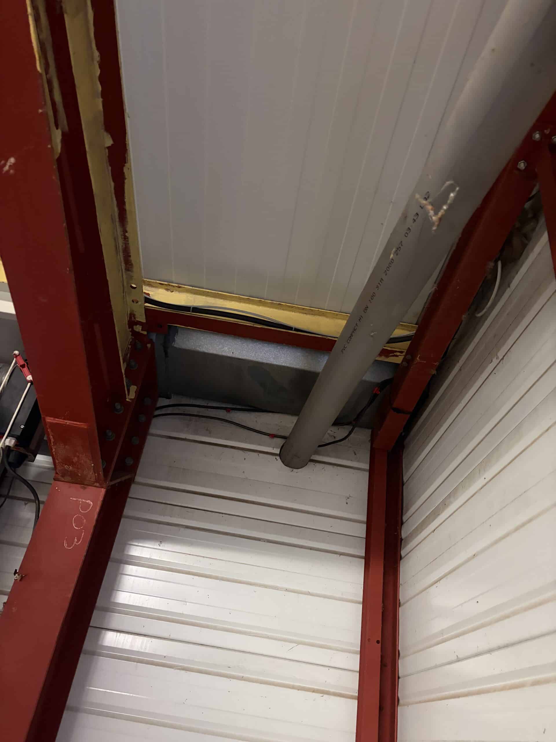 Poutre en acier rouge marquée PO3, tuyau PVC gris et panneaux blancs ondulés dans une structure industrielle.