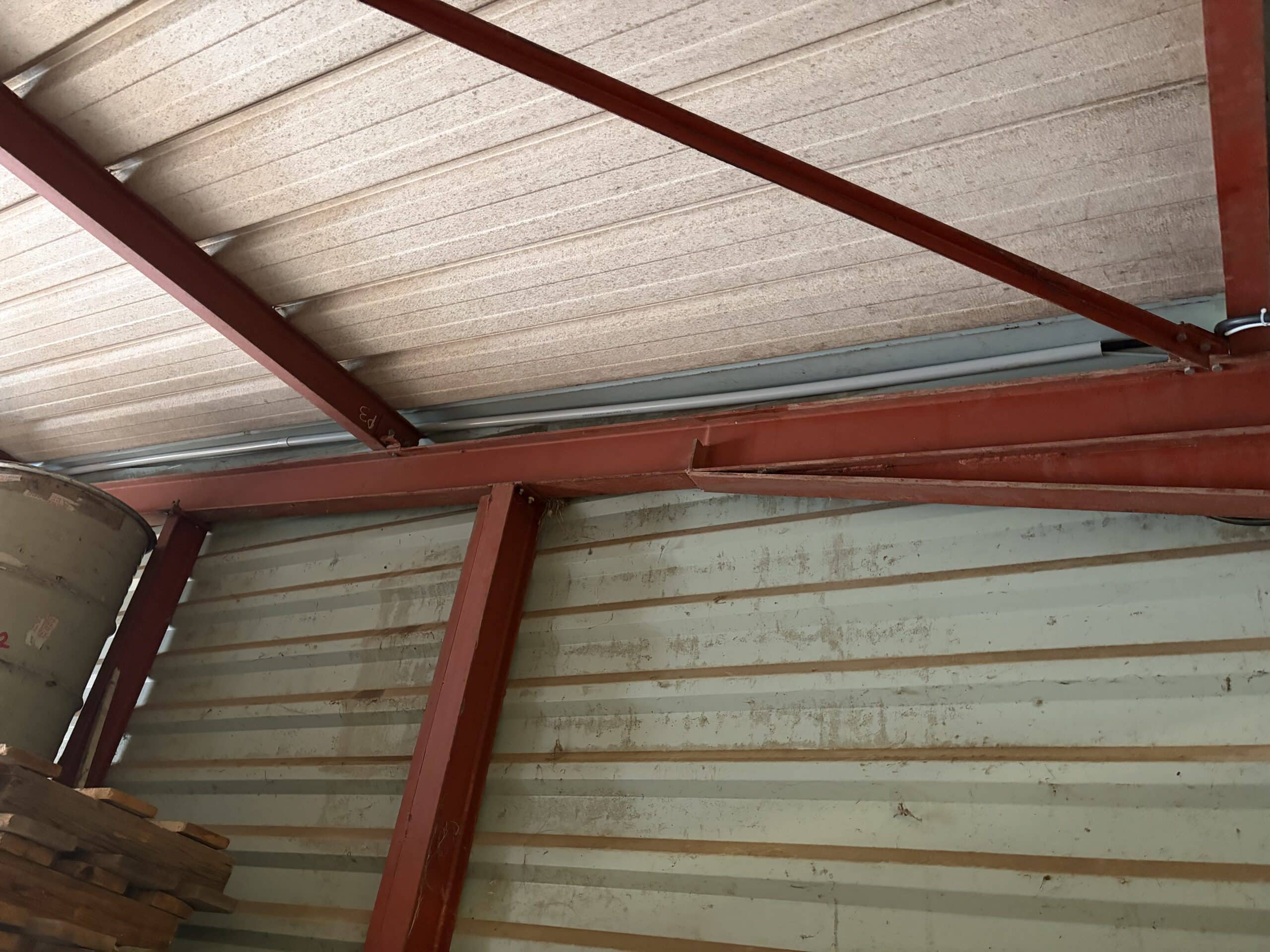 Charpente métallique rouge présentant une poutre pliée sous un toit en tôle ondulée.